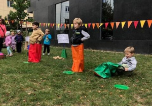 Na zdjęciu widać dwóch chłopców stojących w kolorowych workach. Trzeci chłopiec siedzi na trawie i próbuje włożyć nogi do worka. Za dziećmi rozciągają się kolorowe proporczyki. W tle widać ciemną ścianę budynku.