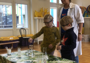 Na stole przykrytym obrusem w zielone liście stoją pojemniki laboratoryjne z różnokolorowymi cieczami. Za stołem stoi chłopiec z dziewczynką i wlewają płyn z menzurki do naczynia. Za nimi stoi kobieta ubrana w biały fartuch.