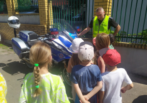 Na zdjęciu widać policjanta stojącego przy policyjnym motocyklu. Przed nim stoją dzieci. W tle widać ogrodzenie