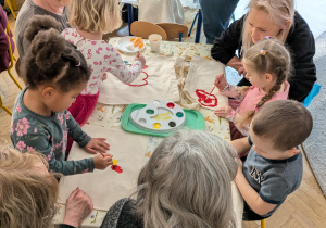 Na zdjęciu widać grupkę dzieci oraz dorosłych, którzy siedzą przy stolikach i malują torby materiałowe.
