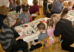 Na zdjęciu widać grupkę dzieci oraz dorosłych, którzy siedzą przy stolikach i malują torby materiałowe.