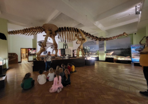 Zdjęcie przedstawia dzieci siedzące tyłem, przed eksponatem szkieletu diplodoka.