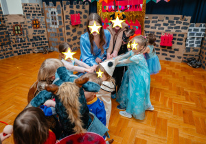 Grupa dzieci w kolorowych strojach karnawałowych, w tym księżniczek, rycerzy i postaci bajkowych, bierze udział w zabawie w udekorowanej sali przypominającej zamek. Dzieci trzymają się za ręce i wspólnie wykonują zadanie, z uśmiechami na twarzach. W tle widać kartonowe dekoracje przypominające mury zamkowe, kolorowe girlandy oraz nauczycielki lub opiekunki pomagające dzieciom.