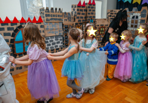 Dzieci ustawione w długi, wesoły pociąg trzymają się za ubrania swoich kolegów i koleżanek, biorąc udział w zabawie. Wystrojone są w bajkowe kostiumy, m.in. księżniczek i astronautów. Sala ozdobiona jest kartonowymi dekoracjami przypominającymi średniowieczny zamek, z kolorowymi wieżyczkami i oknami. Atmosfera jest pełna radości i wspólnej zabawy.