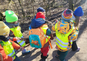 Dzieci w kolorowych kurtkach i kamizelkach odblaskowych niosą gałęzie. W tle widać drzewa.