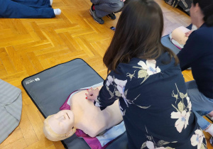 Zdjęcie przedstawia kobietę w kwiecistej bluzce, uciskającą klatkę piersiową fantoma.