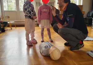 Zdjęcie przedstawia dwie dziewczynki stojące nad fantomem przy instruktorze pierwszej pomocy. Instruktor trzyma telefon komórkowy skierowany w stronę dziewczynek.