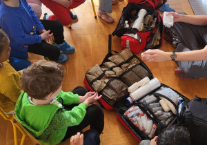 Zdjęcie przedstawia rozłożona torbę ratowniczą pierwszej pomocy wokół której siedzą dzieci na krzesłach