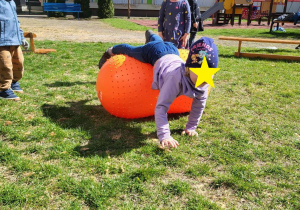 Zdjęcie przedstawia dziewczynkę leżącą na pomarańczowej piłce sensorycznej. W tle widać dzieci obserwujące dziewczynkę.