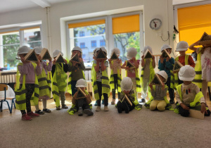 Dzieci w wieku przedszkolnym stojące w białych kaskach i kamizelkach odblaskowych prezentują swoje prace z drewna