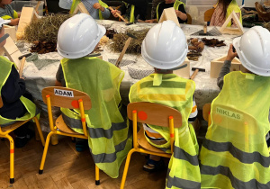Dzieci w kaskach i kamizelkach odblaskowych siedzą przy stole warsztatowym.