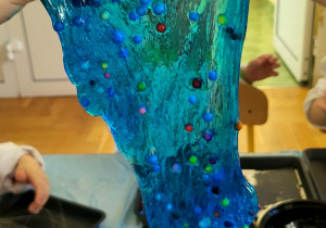 Na zdjęciu widać ręce trzymające niebieski slime z kolorowymi kulkami