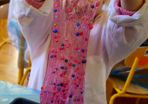 Dziecko w białym fartuchu trzymające przed twarzą różowy slime ściekający do czarnego pojemnika