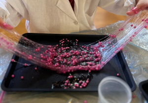 Na zdjęciu widać ręce rozciągające różowy slime znajdujący się w czarnym pojemniku.