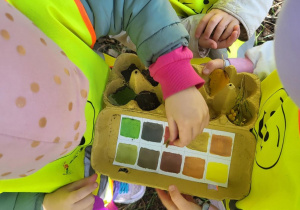 Dzieci w kamizelkach odblaskowych porównują zebrane liście i naturalne materiały z paletą kolorów w kartonowym pudełku.
