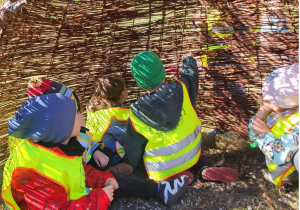 Dzieci w kolorowych czapkach i odblaskowych kamizelkach siedzą wewnątrz wiklinowej konstrukcji na świeżym powietrzu, obserwując otoczenie.