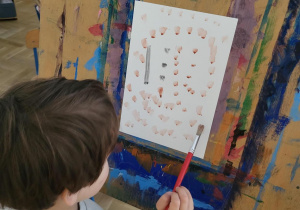 Zdjęcie przedstawia chłopca stojącego przy sztaludze i malującego obraz z kropek.