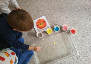 Zdjęcie przedstawia chłopca używającego pipety do zrobienia kolorowej kropki w pojemniku pełnym wody. Wokół pojemnika stoją kubeczki z kolorowym płynem oraz leży książka pt. "Kropka"