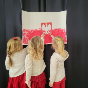 Trzy dziewczynki ubrane w biało-czerwone stroje stoją przed pracą plastyczną przedstawiającą flagę Polski z godłem i wskazują na biało-czerwony orzeł w centralnej części