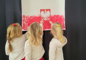 Trzy dziewczynki ubrane w biało-czerwone stroje stoją przed pracą plastyczną przedstawiającą flagę Polski z godłem, wskazują na nią palcem. Na plakacie widnieje biało-czerwony orzeł w centralnej części, tło jest białe na górze i czerwone na dole, wygląda jak flaga Polski.