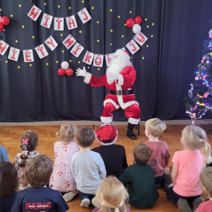 Zdjęcie przedstawia grupę dzieci siedzących na sali i wpatrzonych w postać Świętego Mikołaja i Elfa.