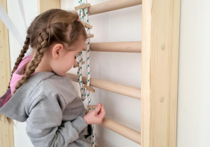 dziewczynka w warkoczach wspięta na drabinkę gimnastyczną przyczepia drewniany spinacz do linki