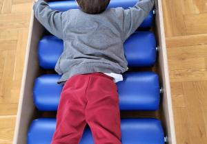 Chłopiec w szarym swetrze i czerwonych spodniach leży na niebieskiej rolce sensorycznym