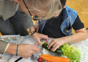 Kobieta krojąca zielonego ogórka którego trzyma dziecko. Obok ogórka na talerzu leży marchewka, rzodkiewka i liście sałaty.