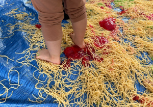 stopy dziecka stojące na niebieskiej plandece pokrytej makaronem spaghetti i różowym kiślem