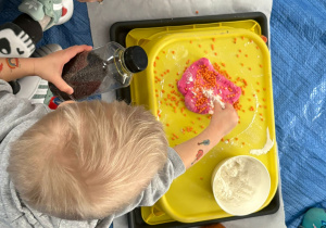 Dziecko pochylone nad tacą posypuje mąką różową masę plastyczną