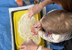 Dziecko i matka pochyleni nad tacą z wysypaną mąką. Dziecko rękoma ugniata różową masę plastyczną