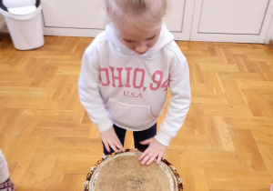 dziewczynka ubrana w białą bluzę z różowym napisem stojąca przy bębnie afrykańskim