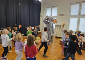 grupa przedszkolaków tańcząca w kole z afrykańskimi instrumentami w rękach wpatrzona w postać słonia, z lewej strony zza dzieci widać głowę ciemnoskórego mężczyzny