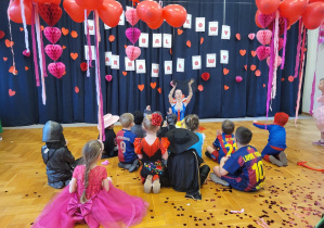 Zdjęcie przedstawia grupke dzieci w przebraniach siedzących przodem do kobiety w przebraniu Królewny Śnieżki. W tle widać ozdoby walentynkowe czerwone balony, serpentyny