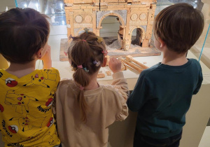Troje dzieci stoi przed gablotą z eksponatem przedstawiającym miniature starożytnego budynku